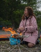 A woman kneels beside a small campfire on a rocky ground, wearing a light pink COLDWATERCO waterproof jacket. A blue bag, folding camp table, and part of an orange sleeping bag are visible in an outdoor wooded setting.