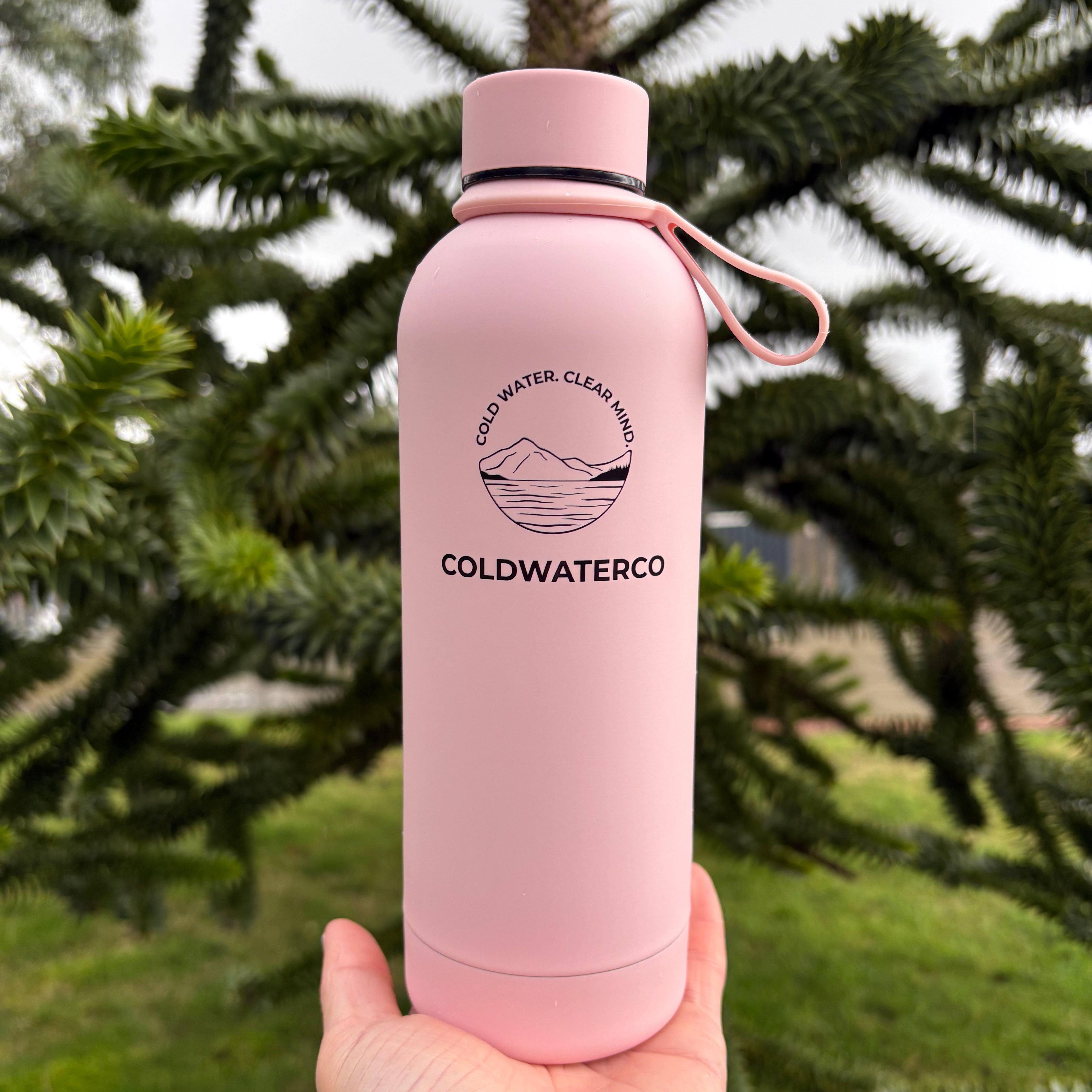 Pink water bottle with 'Coldwater Co.' logo held by a hand against a green natural background