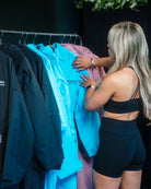 A woman with blonde hair wearing a black sports bra and biker shorts browses a clothing rack filled with jackets in black, light blue, and pink.