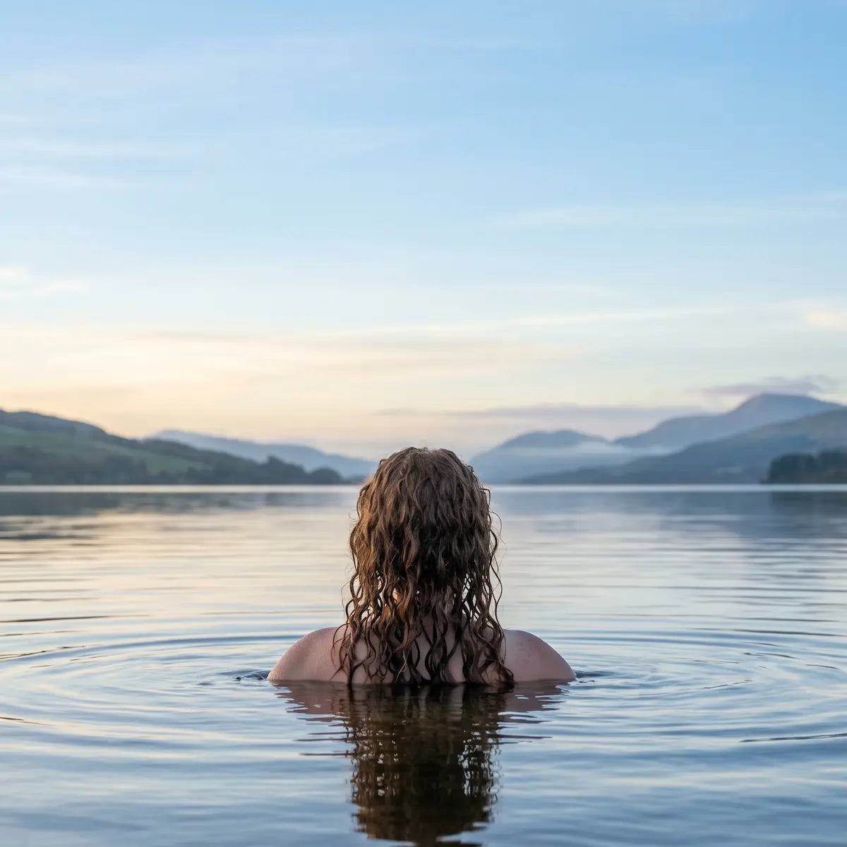 Person with wet hair standing in a lake with mountains in the background