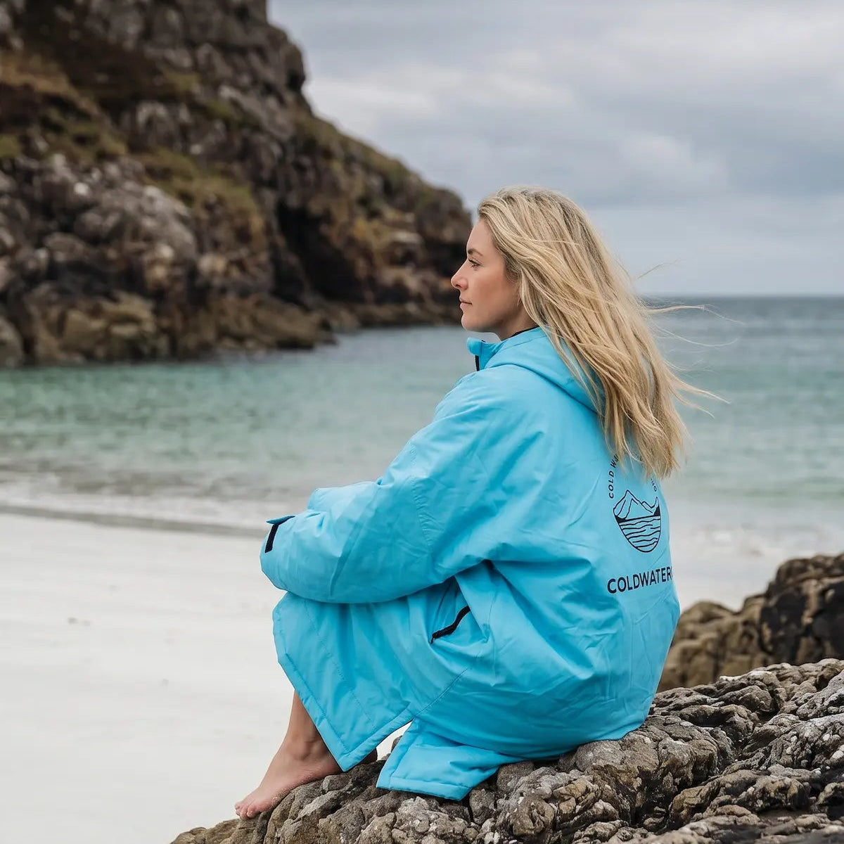 Woman seated on rocks at white beach with ColdWaterCo robe on
